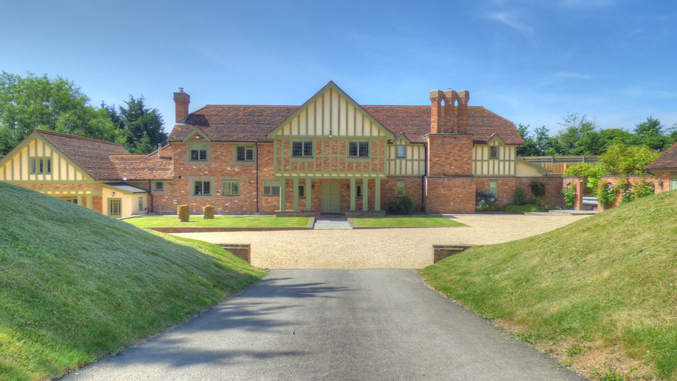 A large, two-story Tudor-style house with brick and timber detailing, surrounded by green lawns and trees, viewed from a driveway leading up to a gravel courtyard—perfect for fully managed holiday lets or Airbnb management.