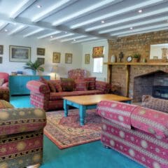 Cozy living room with patterned sofas, a wooden coffee table, brick fireplace, framed art on white walls, potted plants, teal cabinets, and exposed beams—perfect for relaxing after Holiday Home Cleaning or enjoying Fully Managed Holiday Lets.