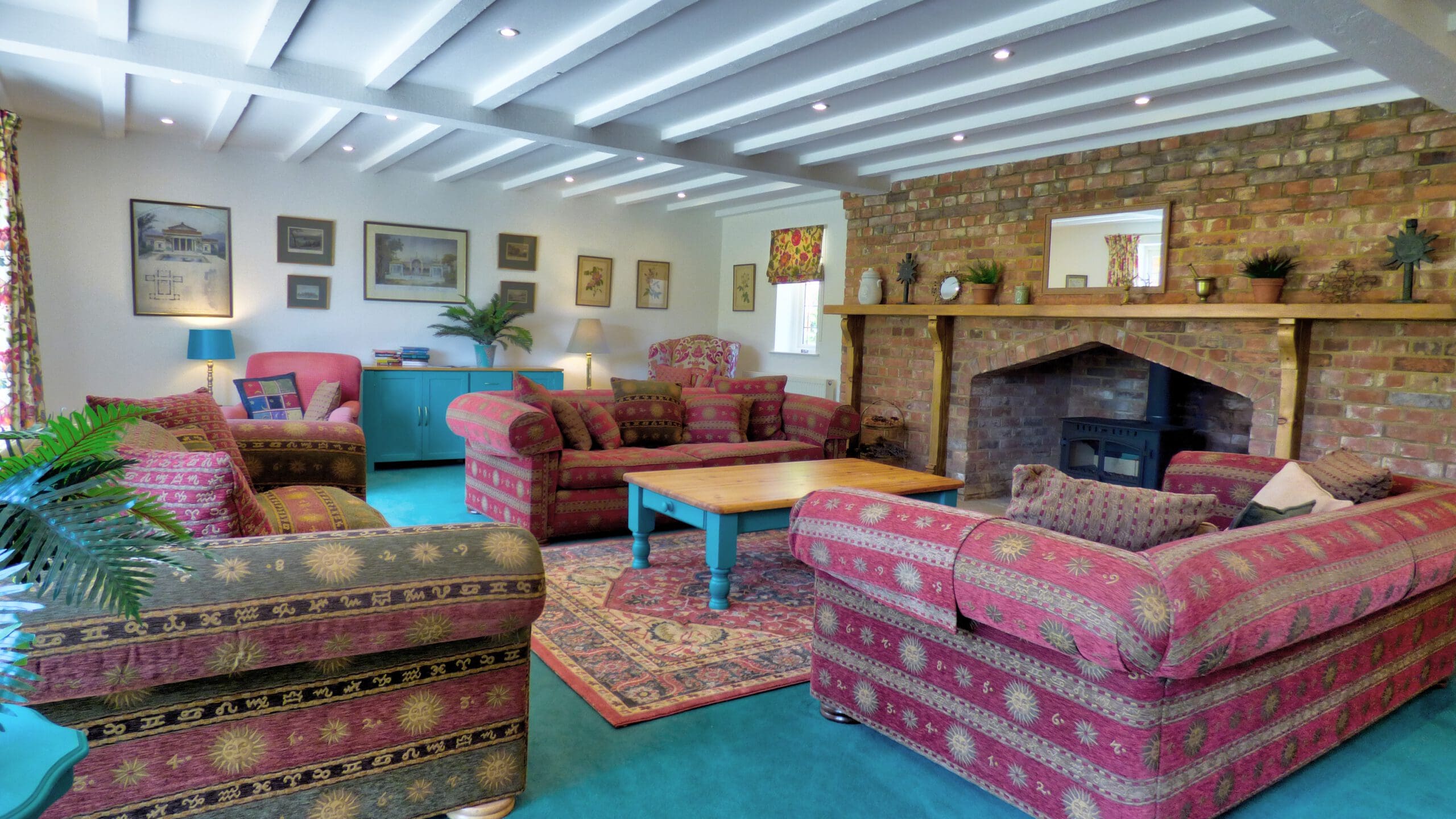 Cozy living room with patterned sofas, a wooden coffee table, brick fireplace, framed art on white walls, potted plants, teal cabinets, and exposed beams—perfect for relaxing after Holiday Home Cleaning or enjoying Fully Managed Holiday Lets.