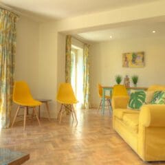 Bright living and dining area with yellow chairs, a yellow sofa with patterned cushions, wooden herringbone floor, large windows, and colorful curtains overlooking a green garden—ideal for effortless property management or second home services.