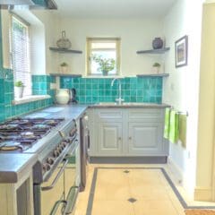 Bright kitchen with turquoise tiled backsplash, gray cabinets, stainless steel gas stove, and a white sink under a window. Perfectly maintained for your property management needs, with green towels and plants adding charm to this inviting space.