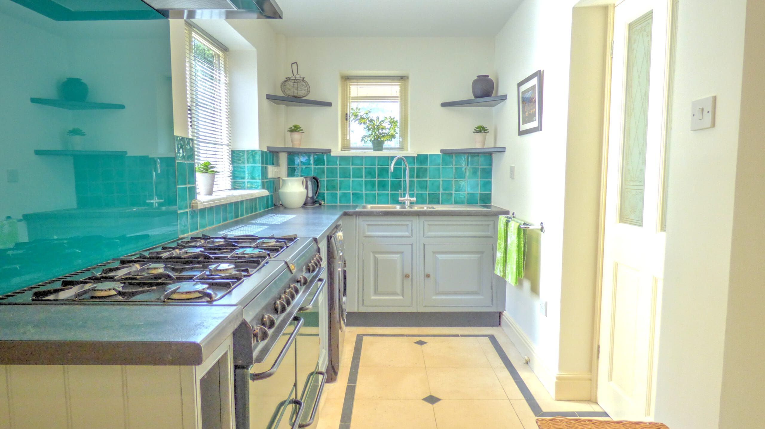 Bright kitchen with turquoise tiled backsplash, gray cabinets, stainless steel gas stove, and a white sink under a window. Perfectly maintained for your property management needs, with green towels and plants adding charm to this inviting space.