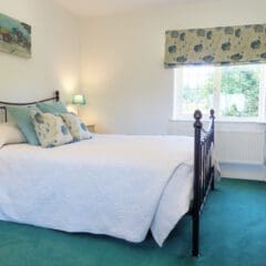 Bright bedroom with a double bed, white bedding, blue and floral accents, nightstands with lamps, a window with floral blinds, a beige armchair, and a vase of white flowers—ideal for clients seeking Second Home Services or Property Management.